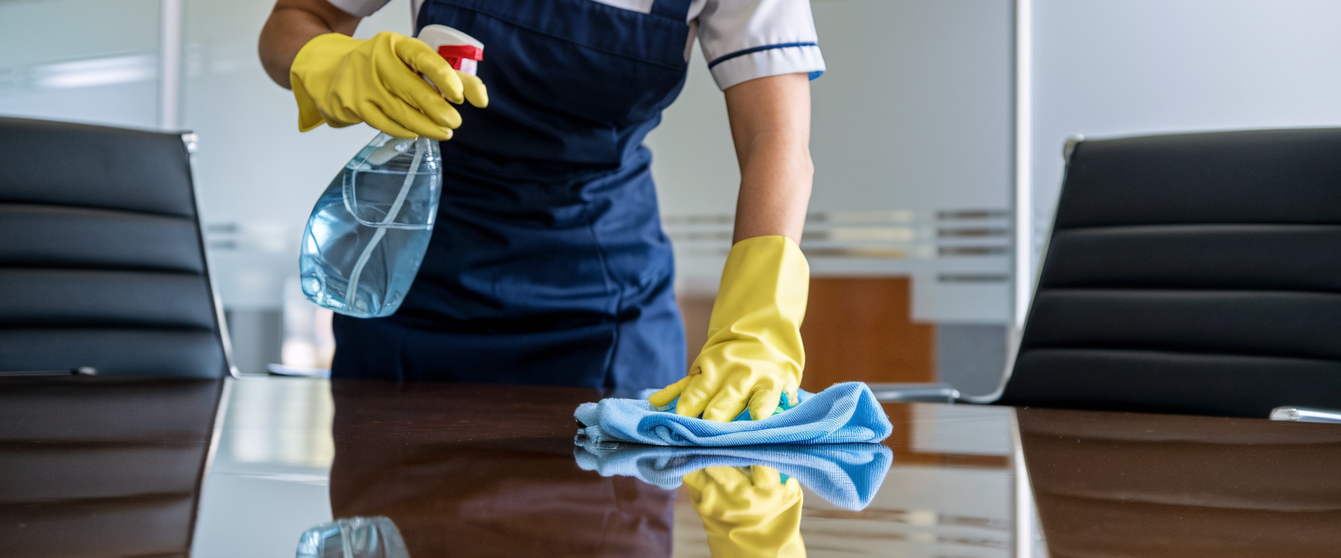 Reinigungspersonal desinfiziert Konferenztisch. Ein engagierter Reinigungsfachmann in blauer Uniform und gelben Handschuhen reinigt sorgfältig einen Glastisch in einer modernen Büroumgebung.