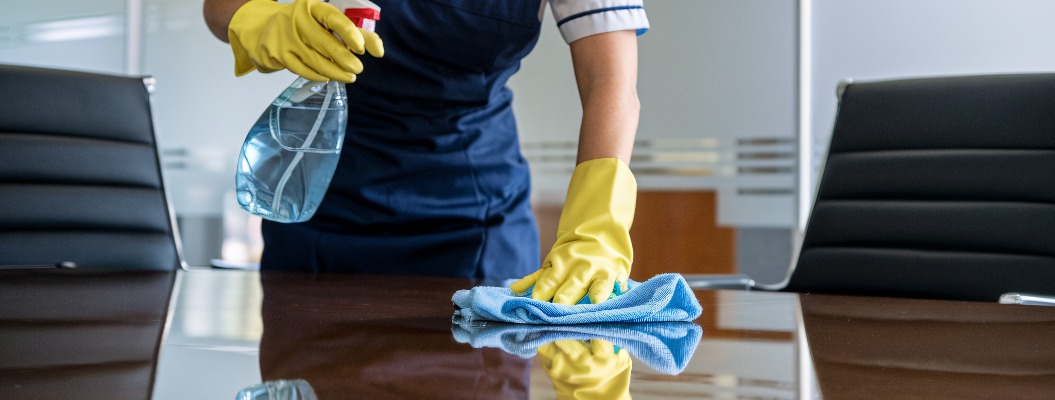 Reinigungspersonal desinfiziert Konferenztisch. Ein engagierter Reinigungsfachmann in blauer Uniform und gelben Handschuhen reinigt sorgfältig einen Glastisch in einer modernen Büroumgebung.